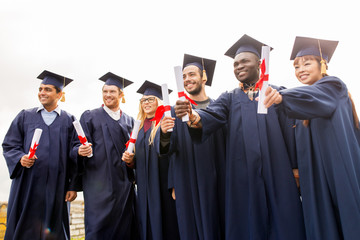 education, graduation and people concept - group of happy international students in mortar boards and bachelor gowns with diplomas