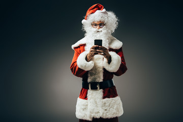 happy santa claus in costume using smartphone isolated on grey background