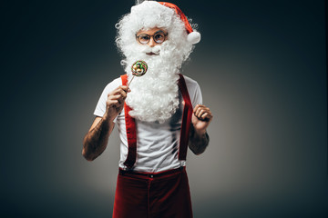 Fototapeta na wymiar santa claus in eyeglasses standing with colorful lollipop isolated on grey background