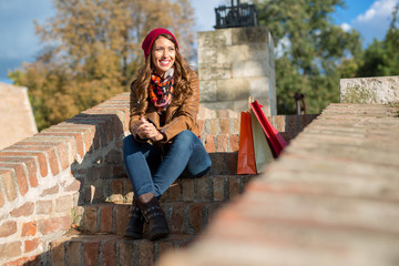 Fototapeta premium Beautiful young woman sitting on stairs outside while drinking a coffee after shopping