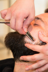 Handsome bearded man is getting shaved by barber at work barbershop