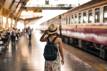 Young woman traveler with backpack waiting for train, Asian backpacker with hat standing on railway platform at Bangkok train station. Holiday, journey, trip and summer Travel concept