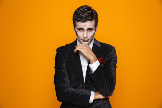 Photo of scary zombie fiance on halloween wearing classical suit and creepy makeup looking at camera, isolated over yellow background
