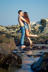 happy couple in love on the beach . beautiful young woman and sexy man hugging and kissing by the sea against the rocks stones , honeymoon trip newlyweds