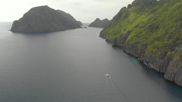 Aerial drone shot following paraw boat driving near limestone cliff island, on a cloudy day, in the archipelago of Palawan, El Nido, Philippines
