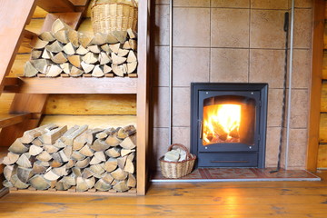 Part of  the room interior in a rural house with a stack of firewood and  a stove with burning wood.