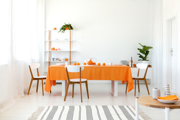 Long dining room table covered with orange tablecloth and comfortable white chairs around it.