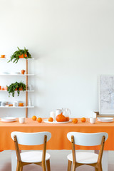Vertical view of dining room table set for dinner. White plates and mugs on orange tablecloth. Shelf with tableware and plants behind it.