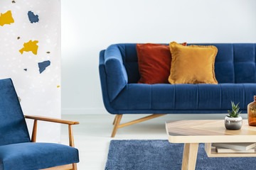 Armchair next to wooden table and blue sofa with pillows in modern living room interior. Real photo