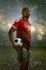 Soccer player on professional soccer night rain stadium. Dirty player in rain drops hold a football ball