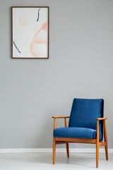 Navy blue wooden armchair in simple grey living room interior with poster on the wall. Real photo