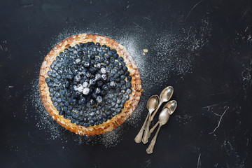 Delicious homemade blueberry almond tart with fresh blueberries and bunch of teaspoons beside. Top  view, blank space, dark toned image - obrazy, fototapety, plakaty