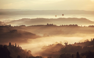 Tuscany at misty morning. Rural landscape in fog during sunrise time and hills, garden trees,...