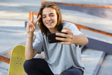 Young skater guy sit in the park with skateboard take selfie by mobile phone with peace gesture. © Drobot Dean