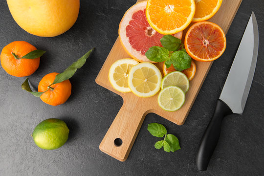 food, healthy eating and vegetarian concept - close up of citrus fruits, wooden cutting board and kitchen knife on slate table top