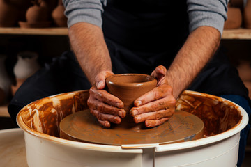 handsome young potter man in a gray sweater and a black apron sculpts clay pot on a potter's wheel in a cozy craft workshop. businessman owner of a dishes china shop