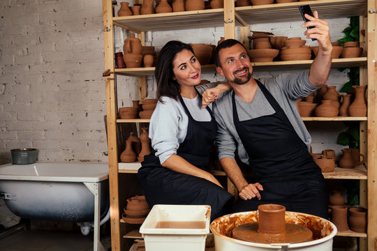 portrait of a beautiful potter woman and happy man doing selfie on the phone. artist family working with clay on a potter's wheel in the pottery workshop. love and art