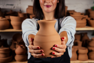 young and attractive female potter working in a studio with a brown clay vase pot in the craft workshop holding the tool and cup in hand . business woman in pottery shop