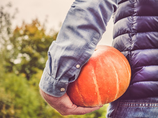 Young man in a denim shirt holding a ripe, yellow pumpkin on a background of green trees. Concept of harvesting and agriculture