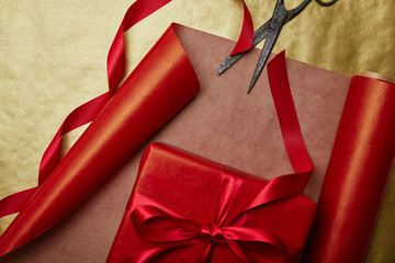 top view of scissors and christmas present with ribbon on red and golden wrapping papers background