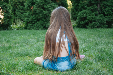 Obraz premium little cute girl with long hair sitting in the park, view from the back on a summer