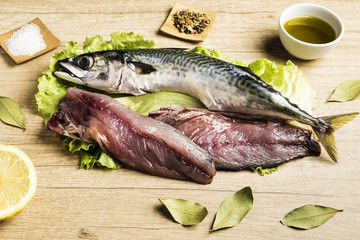Mackerel fresh fish on lettuce leaves next to bay leaves, a few pieces of lemon, a bowl of oil and some spices on a wooden table