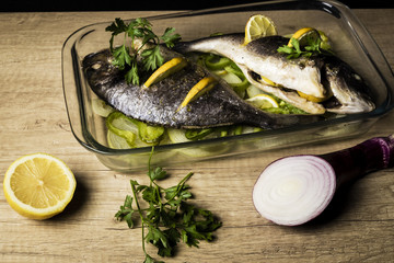 two Dorada fish cooked in the oven with lemon, onion and green pimento in a glass dish on a black slate tray
