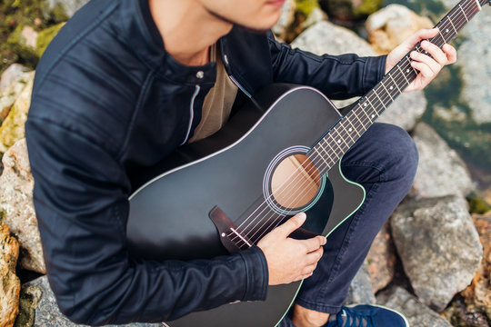 Closeup of young man playing acoustic guitar on beach surrounded with rocks on rainy day