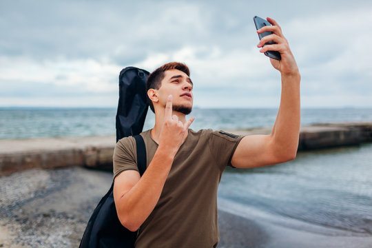 Young man carrying acoustic guitar and taking selfie using phone on cloudy beach. Guy shows rockstar symbol