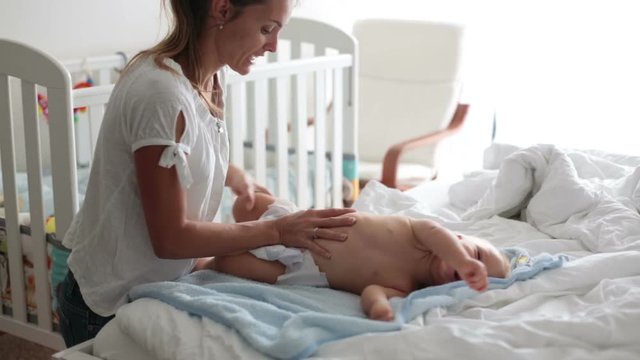 Young mom, changing baby diaper after bath in sunny bedroom