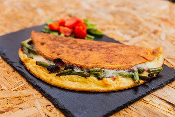 A horizontal photo of omelette with green beans, cheese and mushrooms, garnished with rocket salad and cherry tomatoes. Served on a black stone plate.