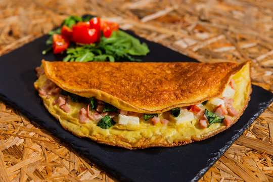 A horizontal photo of omelette with ham, spinach and cheese, garnished with rocket salad and cherry tomatoes. Served on a black stone plate.
