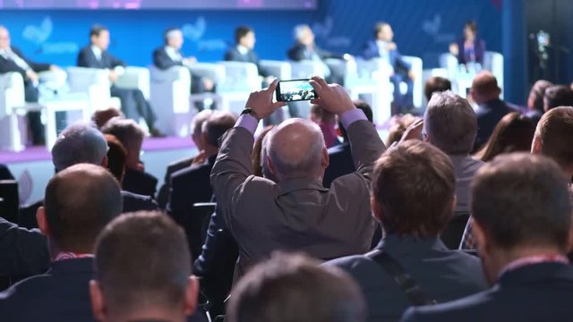 An old businessman or politician is listening to a seminar or lecture on the economic situation. Makes photos or shoots video on a smartphone