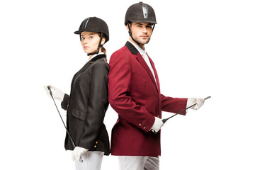 serious young equestrians in uniform and helmets holding horseman sticks while leaning back to back and looking at camera isolated on white
