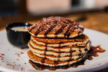 A horizontal photo of pancakes topped with chocolate sauce, powdered sugar and chopped chocolate, served on a white plate with sweetened condensed milk