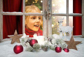 Kind schaut freudig überrascht nach rechts aus dem weihnachtlich dekorierten Weihnachtsfenster,...