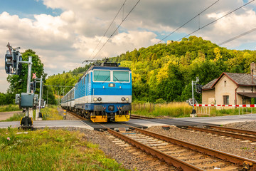 The blue electric train arriving at the crossing with the barriers. A train running through the green valley. Rail transport in the Czech Republic. A sunny day on the railroad