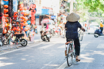Woman in a traditional rice hat riding a bicycle down the streets of Hanoi, Vietnam. Street traffic...
