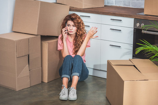 irritated woman with curly hair sitting on floor near cardboard boxes, talking by smartphone and gesturing at new kitchen