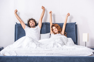 happy young couple stretching arms and waking up together in bedroom - obrazy, fototapety, plakaty