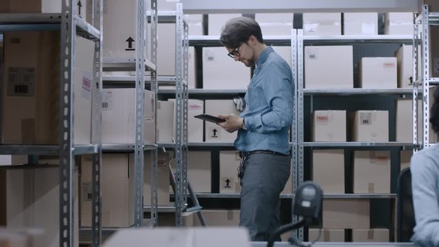 Warehouse Worker Uses Digital Tablet Computer to Checks Stock on Shelves For the Right Cardboard Box Package. In the Background His Female Colleague Works on a Personal Computer.