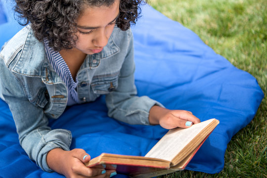 African American girl lying on a blue blanket reading a book outside on a grassy lawn