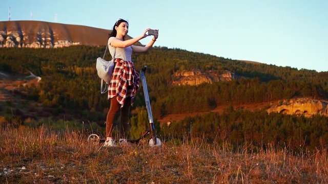 Girl stands next to the electric scooter taking pictures of the mountain landscape and sunset,rear view slow mo