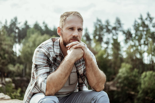 Mountains and trees. Bearded man feeling relieved and truly relaxed while sitting on his own surrounded by mountains and trees