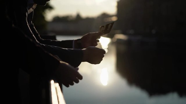 Man and woman watching photos at city embankment. Looking cards at their dating in a town near river at sunrise or sunset silhouette slowmotion