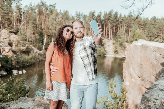 Selfie with girlfriend. Smiling blonde-haired man holding his blue smartphone making selfie with his appealing girlfriend near lake