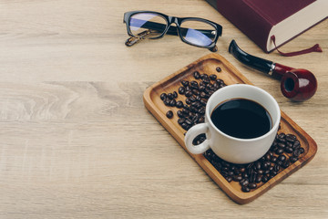 a cup of coffee and a pile of coffee beans in the wooden tray, a red book, a tobacco smoking pipe...