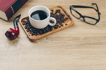 a cup of coffee and a pile of coffee beans in the wooden tray, a red book, a tobacco smoking pipe and an eyeglasses on the wooden table.