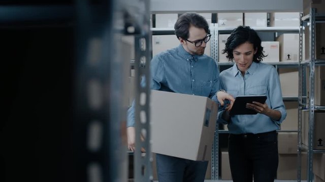 Female Inventory Manager Shows Digital Tablet Information to a Worker Holding Cardboard Box, They Talk and Do Work. In the Background Stock of Parcels with Products Ready for Shipment.