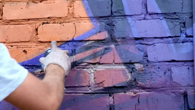 A young guy with a can of paint draws graffiti on the wall. One hand close-up. Slow motion